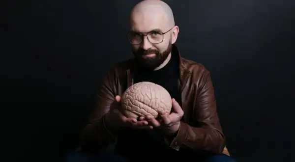 Łysy mężczyzna w okularach i brązowej kurtce trzyma w dłoniach model mózgu.