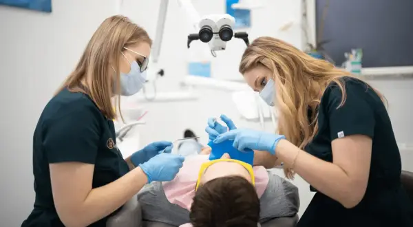 Dwie dentystki w maseczkach i rękawiczkach wykonują dłutowanie zęba u pacjenta pod mikroskopem.