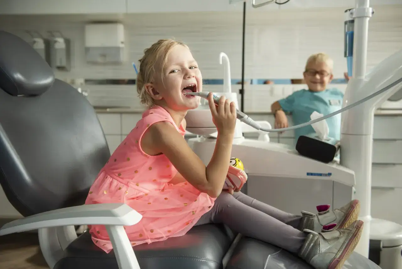 Uśmiechnięta dziewczynka w różowej sukience bawi się wiertłem dentystycznym, gdy dziecko boli ząb mleczak. Obok siedzi chłopiec.
