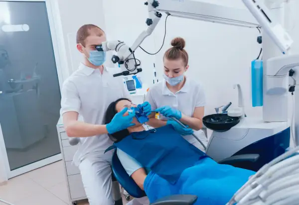 Dentysta pod mikroskopem wykonuje ekstyrpację miazgi u pacjentki. Asystentka pomaga.