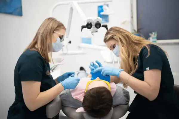 Dwie dentystki w maseczkach i rękawiczkach wykonują dłutowanie zęba u pacjenta pod mikroskopem.